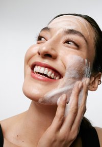 Aanbrengen van gezichtsreiniger op het gezicht van een glimlachende vrouw, met een schuimende textuur, een gladde huid en een schone, lichtgekleurde achtergrond.