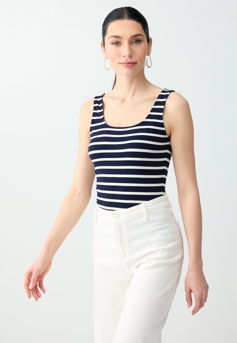 Femme aux cheveux foncés tirés en arrière, portant des boucles d'oreilles anneaux, un débardeur rayé bleu marine et blanc et un pantalon blanc taille haute, debout devant un fond blanc.