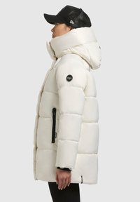 Fehér párnázott dzseki magas gallérral és nagy kapucnival, cipzáras zsebből, fekete fém részletekkel és fényes felülettel.