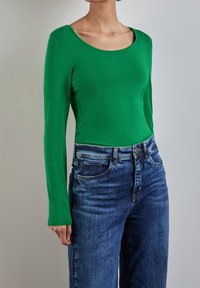 Långärmad grön topp i stretchtyg, parat med högmidjade mörkblå jeans. Toppens har en rund halsringning.