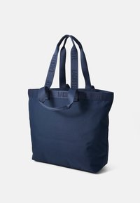 Mørkeblå tote bag laget av mykt stoff med to lange håndtak. Har "LACOSTE"-logo på håndtakene og et romslig, minimalistisk design.