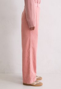 Jasnoróżowe spodnie sztruksowe o prostym kroju, z pionowymi prążkami i kieszeniami po bokach, zestawione z beżowo-brązowymi trampkami.