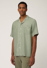 Camisa de manga corta en verde claro, hecha de un material suave. Presenta un bolsillo y un pequeño detalle del logo. Botones en la parte delantera.