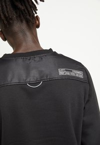 Černá mikina s lesklým saténovým panelem u límce, smyčkovým detailem a tištěnými čísly šarže/referencí na zádech.