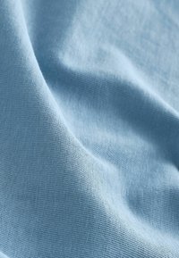Sileä vaaleansininen kangas, jossa hieno neulostekstuuri ja lempeät laskokset luovat pehmeitä varjoja ja korostuksia.