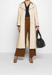 Manteau long beige avec un grand col, boutons dorés, et haut noir ; pantalon marron à jambes larges ; sac à main noir froncé ; talons hauts noirs à lanières.