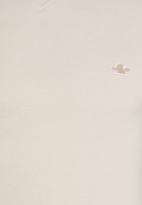 Suéter beige de cuello en v con textura fina de canalé, que presenta un pequeño logo de nube bordado en marrón claro en la parte izquierda del pecho.