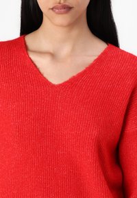 Suéter rojo de punto acanalado con escote en V. Tejido texturizado y bordes ligeramente deshilachados en el escote. Apariencia suave y diseño informal.