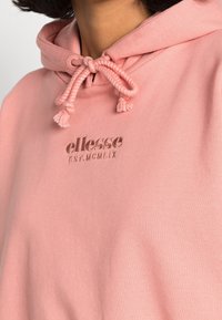 Růžová mikina s kapucí vyrobená z měkkého materiálu, s klokaní kapsou, elastickými šňůrkami a vyšitým logem na hrudi.