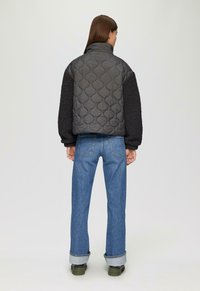 Vrouw met haar rug naar de camera gekeerd, gekleed in een zwarte gewatteerde jas met textuur op de mouwen, blauwe opgerolde jeans en groene schoenen, tegen een effen lichte achtergrond.