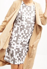 Tumma pitkä takki valkoisen mekon päällä, jossa on harmaa abstrakti kuvio. Mekossa on pyöreä kaula-aukko ja rento leikkaus, sekä lyhyet hihat.