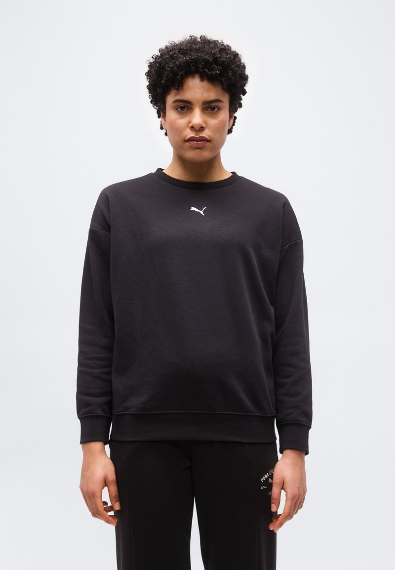 Άτομο με κοντά σγουρά μαλλιά, φορώντας μαύρο φούτερ Puma και μαύρο παντελόνι, στέκεται μπροστά σε απλό φωτεινό φόντο.