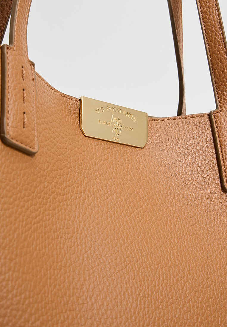 Sac fourre-tout en cuir tan avec une surface texturée et des accents en métal doré. Présente une plaque rectangulaire avec le logo de la marque. Design épuré.