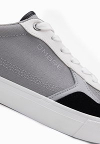 Šedé plátěné tenisky s černými semišovými akcenty, bílou gumovou podešví a silnými bílými tkaničkami s logem vzorovaným na boku.