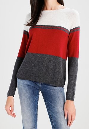 Frau trägt einen langärmeligen Pullover mit weißen, roten und dunkelgrauen horizontalen Farbblöcken, kombiniert mit hellblauen Jeans.