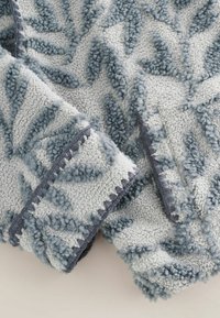 Manta de forro polar suave y texturizada con patrones elevados de hojas azules sobre un fondo gris claro, con bordes cosidos.