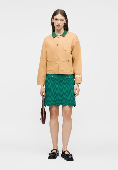 Γυναίκα με ταμπά σακάκι με κουμπιά και πράσινο γιακά, πράσινη φούστα με κυματιστό κόψιμο, μαύρα παπούτσια, κρατώντας μικρή καφέ τσάντα στο πλάι της.