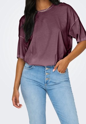 Kobieta w luźnym bordowym t-shircie i niebieskich jeansach z wysokim stanem oraz guzikami z przodu, jedna ręka w kieszeni, na jednolitym tle.