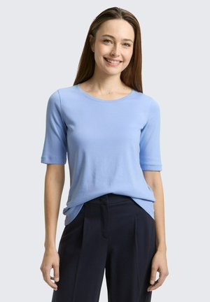 Young woman smiling, wearing a light blue short-sleeve top and dark high-waisted trousers, standing against a plain light background.