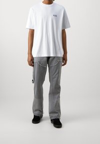 Person iført en hvid kortærmet t-shirt med lille tekst på brystet, grå cargo-bukser med frynsede kanter og sorte sneakers.