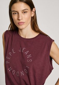 Burgundské bezrukávové tričko vyrobené z měkké tkaniny, s kulatým designem a nápisem "PEPE JEANS LONDON" posetým kamínky na přední straně.