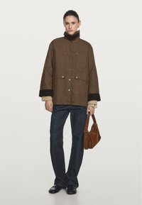 Braune oversized Jacke mit hohem Kragen, Druckknöpfen und zwei vorderen Taschen. Kombiniert mit dunklen Jeans und einer kleinen braunen Tasche.