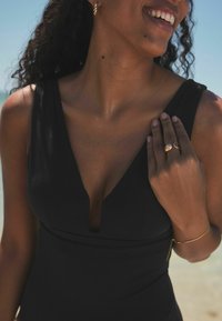 Zwart zwemkleding met een diepe V-hals ontwerp, zichtbaar met een soepele textuur. Gouden sieradenaccenten op hand en oor, weergegeven tegen een strandachtergrond.