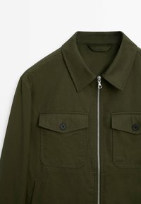 Veste vert olive avec un col pointu, fermeture éclair centrale argentée et deux poches avant à rabat boutonné.