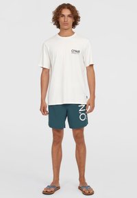 T-shirt en coton blanc avec un logo noir sur la poitrine, associé à un short de bain teal avec une branding blanche, et des sandales marron avec des bretelles bleues.