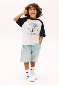 Χαμογελαστό παιδί με σγουρά μαλλιά, που φοράει ένα δίχρωμο T-shirt Tommy, ανοιχτόχρωμα τζιν σορτς και λευκά αθλητικά παπούτσια με κάλτσες επωνυμίας, στέκεται με το ένα χέρι στην τσέπη.