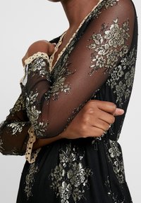 Vestido negro de malla con bordado floral dorado, con puntilla en las mangas y cintura ajustada. Presenta patrones intrincados y una textura transparente.