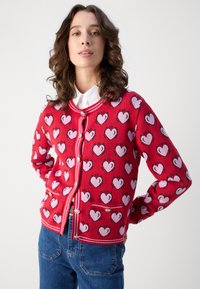 Punainen neuletakki, jossa on vaaleanpunainen sydänkuvio, poimutettu kaulus ja hihansuut, etupainikkeet ja pieni tasku. Päällä valkoinen kauluspaita.