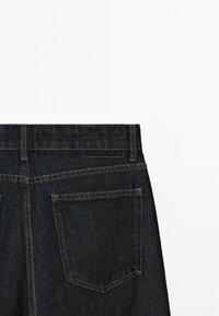 Donkerdenim jeans met een hoge tailleband, twee achterzakken en contrasterende oranje stiksels langs de naden. Textuur en stevig materiaal.