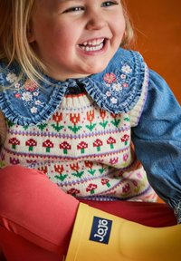 En skrattande småbarn som har på sig en blommigt broderad denimkrage, en mönstrad stickad tröja, röda leggings och gula gummistövlar.