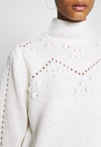 Vit stickad tröja med hög ribbad krage, med strukturerade boblemönster och öppna stygn detaljer längs axelsömmarna.