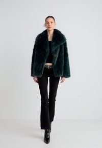 Chaqueta de piel sintética en color azul verdoso oscuro con un amplio cuello tipo chal. La modelo lleva un top corto negro y jeans acampanados, completando el look con botas negras brillantes.