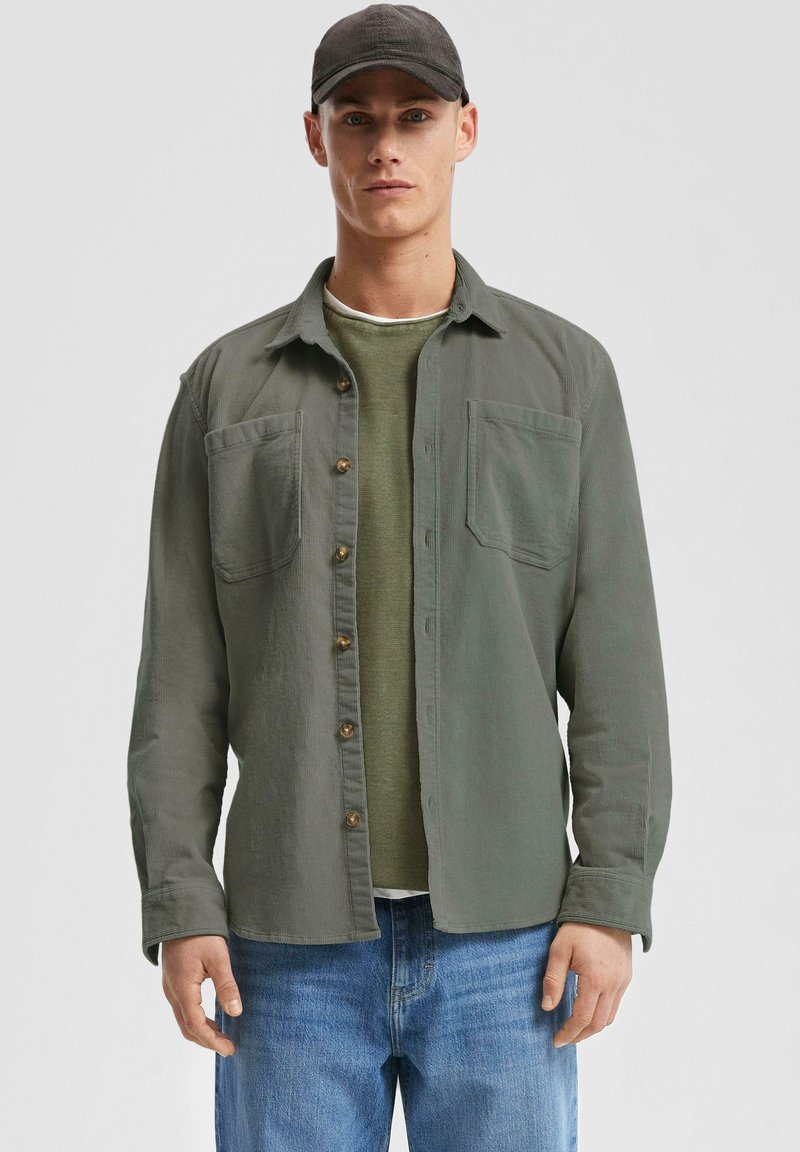 Groene button-up shirt met twee borstzakken, gedragen over een lichtgroen overhemd met lange mouwen. Model draagt een donkere pet en lichtblauwe jeans.