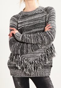 Czarnoskalisty sweter z przędzy w stylu melanżowym z frędzlami, noszony z rękami skrzyżowanymi na piersi.