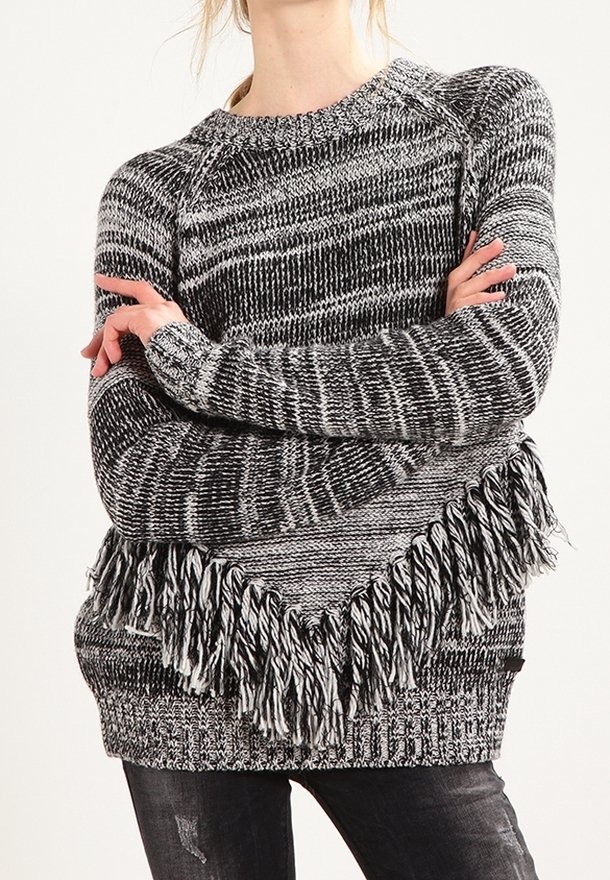 Czarnoskalisty sweter z przędzy w stylu melanżowym z frędzlami, noszony z rękami skrzyżowanymi na piersi.