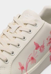 Άσπρο αθλητικό παπούτσι με κρέμ κορδόνια που διαθέτει ροζ λουλουδένια σχέδια και λεπτομέρεια πεταλούδας στο πλάι, κοντινή λήψη στα οπές και τη ραφή.