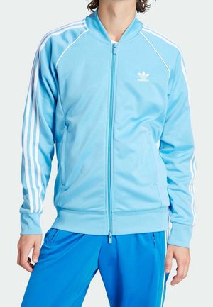 Hombre con chaqueta deportiva Adidas azul claro con franjas blancas en las mangas y pantalones deportivos azules, de pie con las manos relajadas.
