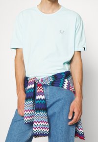 Světle modré bavlněné tričko s krátkými rukávy, na kterém je malé šedé logo. Kolem pasu je uvázaný šátek s vícebarevným cik-cak vzorem.