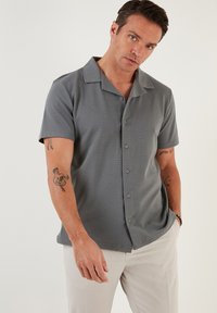 Camisa de botón de manga corta en gris con una textura única, que presenta un cuello plano y sutiles rayas horizontales.