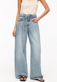 Mujer que lleva unos pantalones vaqueros de tiro alto y pernera ancha de color azul claro, con un cinturón delgado, una blusa sin mangas de color beige y sandalias planas negras.