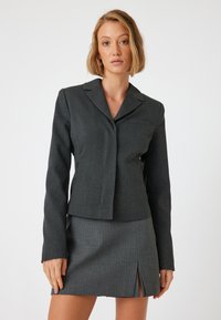 Dunkelgrau maßgeschneiderte Jacke mit Reverskragen, seitlichen Taschen und einem einzelnen Knopfverschluss. Kombiniert mit einem passenden, gestreiften Minirock.