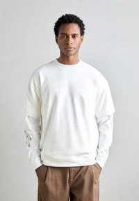 Bílý oversized sweatshirt s kulatým výstřihem, vrstvený přes dlouhý rukáv tričko s grafikou bezpečnostní špendlík na rukávu. Hnědé kalhoty.