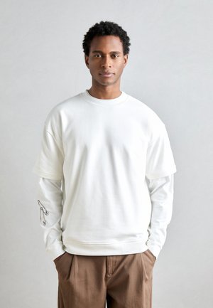 Sweatshirt blanc surdimensionné à col rond, superposé sur une chemise à manches longues avec un graphique d'épingle de sécurité sur la manche. Pantalons marron.