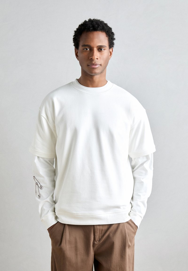 Bílý oversized sweatshirt s kulatým výstřihem, vrstvený přes dlouhý rukáv tričko s grafikou bezpečnostní špendlík na rukávu. Hnědé kalhoty.