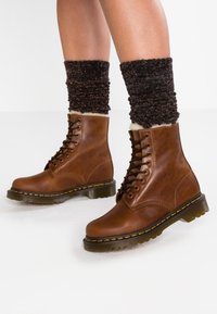 Botas de cuero marrón hasta el tobillo con cordones negros, costuras amarillas y una suela transparente gruesa. Medias de lana oscuras y brillantes visibles sobre las botas.