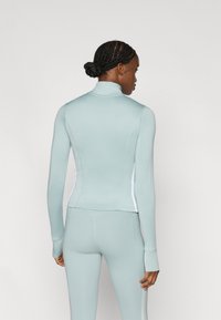 Ένα εφαρμοστό αθλητικό μπουφάν σε ανοιχτό μπλε χρώμα με ψηλό γιακά και μακριά μανίκια, με λευκές πλαϊνές πινελιές για πρόσθετη αντίθεση και απαλή υφή.